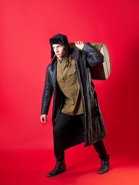 Un hombre con ropa pasada de moda y un sombrero con orejeras, arrastra su gran maleta sobre sí mismo, al estilo ruso de posguerra. Retrato sobre un fondo rojo