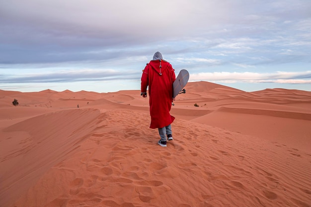 Hombre con ropa tradicional con sandboard caminando sobre dunas de arena contra el cielo