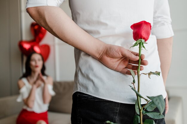 Hombre con rosa en la mano haciendo sorpresa para mujer sentada en el sofá con globos en forma de corazón en casa