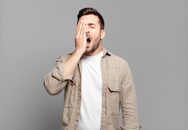 Hombre rubio adulto guapo que parece somnoliento, aburrido y bostezando, con dolor de cabeza y una mano cubriendo la mitad de la cara