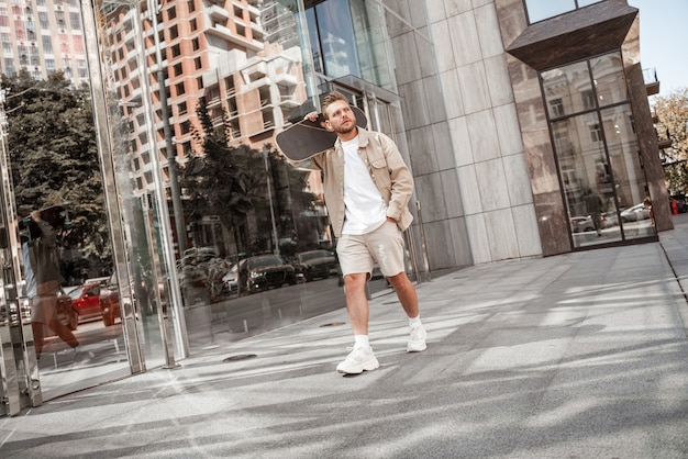 Hombre rubio alegre joven que sostiene el longboard en el hombro en la calle de la ciudad. Viste traje de mezclilla beige. Skater deportivo pensando en un nuevo truco extremo en el exterior.