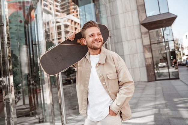 Hombre rubio alegre joven que sostiene el longboard en el hombro en la calle de la ciudad. Viste traje de mezclilla beige. Skater deportivo pensando en un nuevo truco extremo en el exterior.