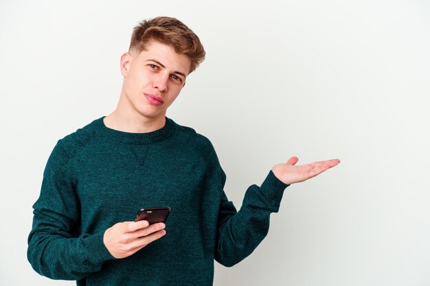 Hombre rubio caucásico joven que usa un teléfono aislado en la pared blanca que muestra un espacio de la copia en una palma y que sostiene otra mano en la cintura.