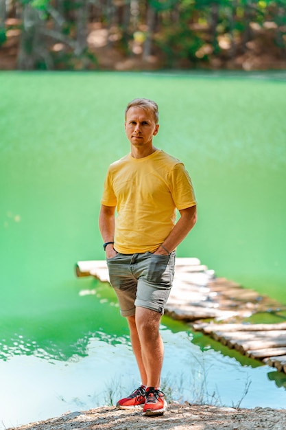 Un hombre rubio está parado frente a un lago de montaña azul en el bosque en verano