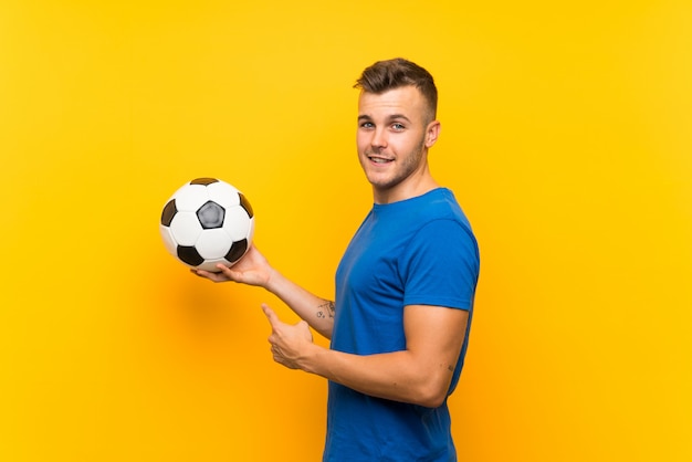 Hombre rubio hermoso joven que sostiene un balón de fútbol sobre la pared amarilla aislada y que lo señala