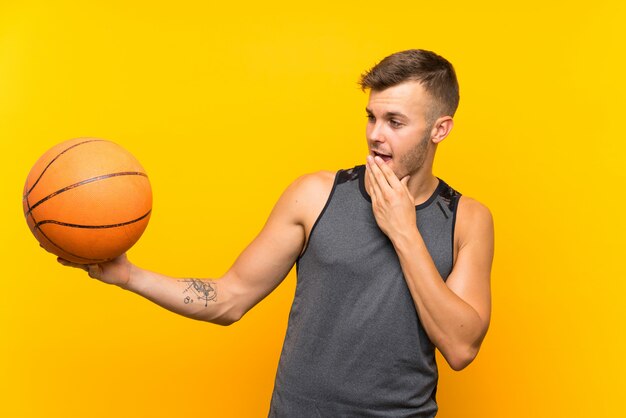 Hombre rubio hermoso joven que sostiene una bola de cesta sobre la pared amarilla aislada con la expresión facial sorprendida y conmocionada