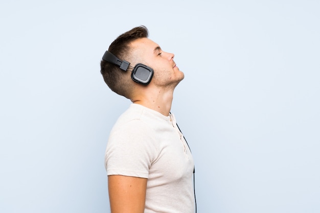 Hombre rubio hermoso joven sobre la pared azul aislada que escucha la música con los auriculares