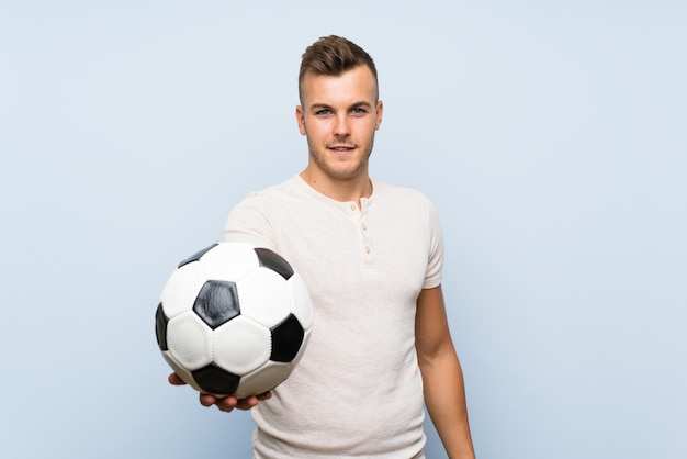 Hombre rubio hermoso joven sobre la pared azul aislada que sostiene un balón de fútbol