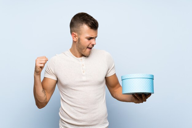 Foto hombre rubio hermoso joven sobre la pared azul aislada que sostiene la caja de regalo