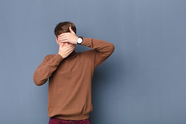 Hombre rubio joven que cubre la cara con ambas manos diciendo que no! rechazar fotografías o prohibir fotografías aisladas contra una pared plana