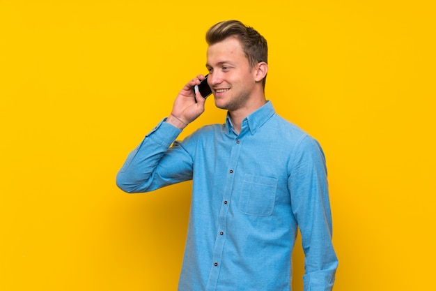 Hombre rubio sobre la pared amarilla aislada que mantiene una conversación con el teléfono móvil
