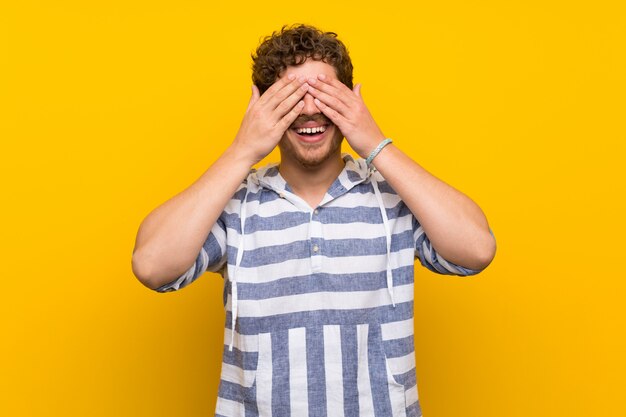 Foto hombre rubio sobre pared amarilla que cubre ojos por las manos. sorprendido de ver lo que viene por delante.