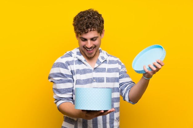 Foto hombre rubio sobre la pared amarilla que sostiene un regalo en manos