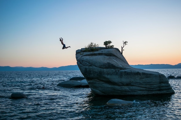 Hombre saltando de una roca por el mar contra un cielo despejado