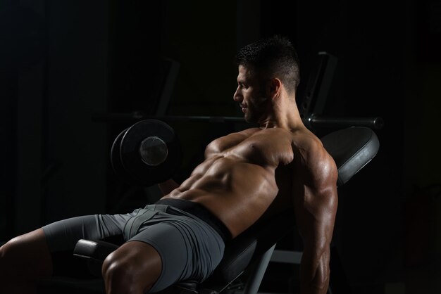 Hombre sano trabajando bíceps en un gimnasio oscuro rizos de concentración con mancuernas