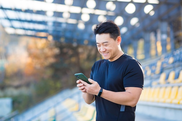 Un hombre sano ve un video en un teléfono móvil después de hacer ejercicio mientras está sentado al aire libre usando una aplicación en un dispositivo inalámbrico 4g El atleta asiático está feliz y sonriendo después de descansar