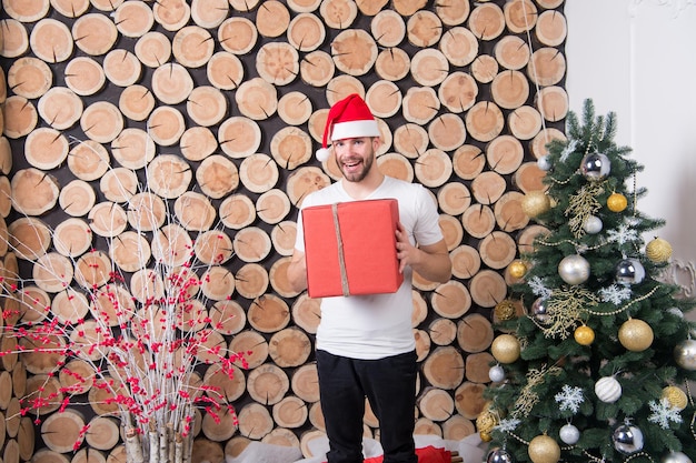 Hombre de Santa con caja en el árbol de Navidad. Macho con sombrero rojo sonrisa con regalo sobre fondo de troncos de madera. Preparación de regalos de navidad y año nuevo. Celebración de felices fiestas. Concepto del día de San Esteban.