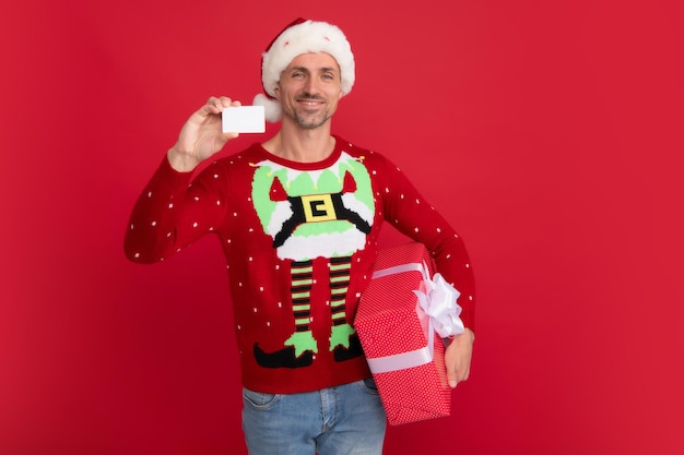 El hombre de Santa sostiene una caja de regalo roja y una tarjeta de crédito Retrato de estudio de un chico que usa un gorro de Papá Noel y un suéter de invierno Hombre con gorro de Navidad sobre un fondo rojo aislado