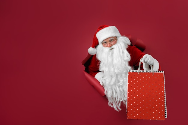 El hombre de Santa sostiene el paquete de tiendas en la mano a través de un agujero de papel. Hombre barbudo con sombrero de santa mirando a través del agujero en papel rojo.