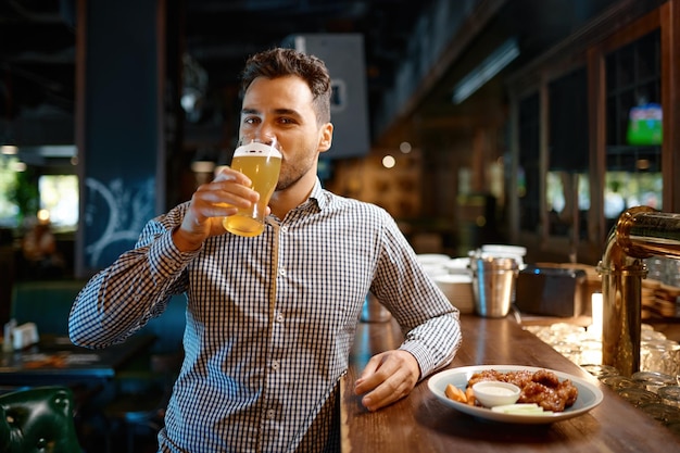 Hombre satisfecho bebiendo deliciosa cerveza artesanal fría y fresca mientras descansa en el bar deportivo. Joven parado en el mostrador mirando a la cámara mientras prueba una bebida espumosa