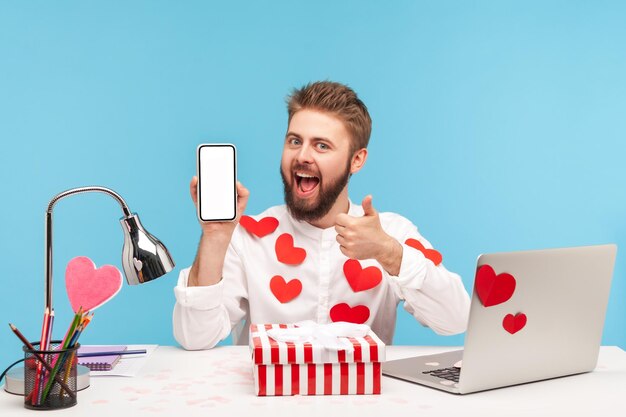 Hombre satisfecho sonriente con barba en pantalones blanco cubierto de corazones pegajosos mostrando los pulgares hacia arriba sosteniendo el teléfono inteligente con pantalla vacía. Disparo de estudio interior aislado sobre fondo azul.