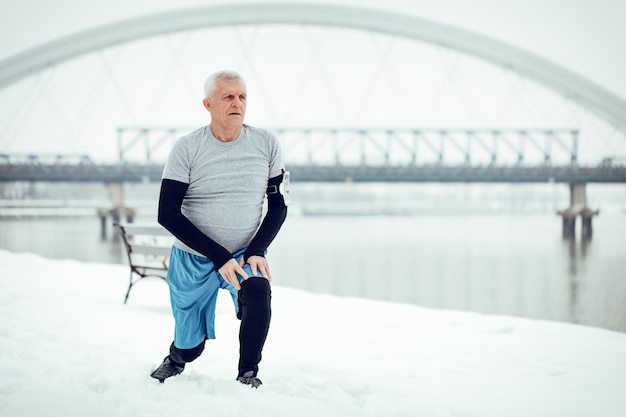 Hombre senior activo estirándose y haciendo ejercicios junto al río durante el entrenamiento de invierno afuera. Copie el espacio.