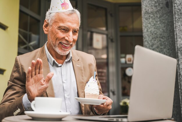 Hombre senior alegre celebrando cumpleaños con videollamada en la terraza del café