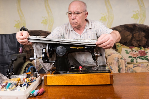 Hombre senior en la antigua máquina de coser midiendo las piernas del pantalón en casa en la sala de estar