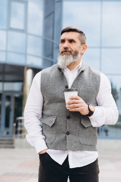 Hombre senior canoso barbudo brutal positivo tomando café en el espacio urbano