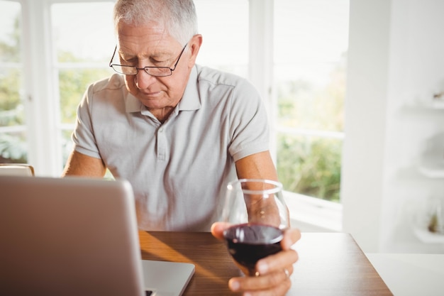 Hombre senior enfocado usando laptop y bebiendo vino en casa