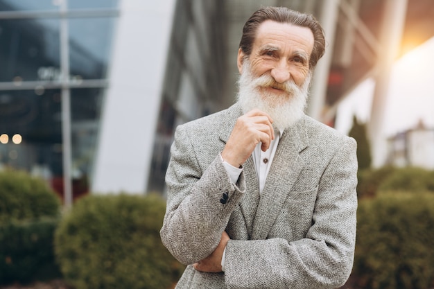 Hombre senior feliz sosteniendo su barba gris en los edificios de oficinas con espacio de copia