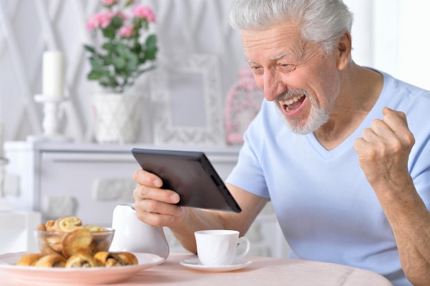 Hombre senior feliz usando tableta mientras bebe té en la cocina