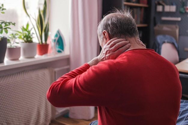 Hombre senior frustrado en suéter rojo con la mano en el cuello sentado en el sofá