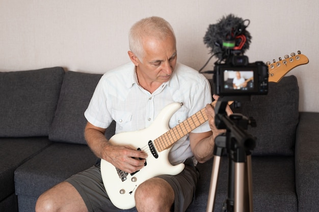 Hombre Senior grabando blog de videos musicales, tocando la guitarra sentado en el sofá en casa