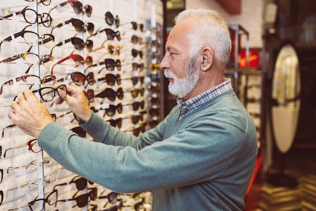 Hombre senior guapo elegir marco de anteojos en tienda óptica.