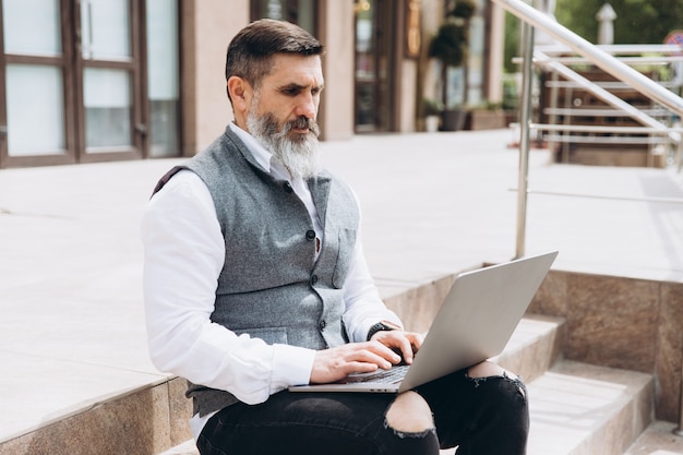 Hombre senior hombre elegante con barba gris pasa tiempo al aire libre en la computadora portátil