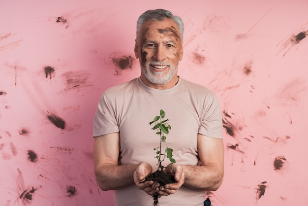 Hombre senior, jardinero sosteniendo una planta para plantar en una pared de pared rosada y sucia