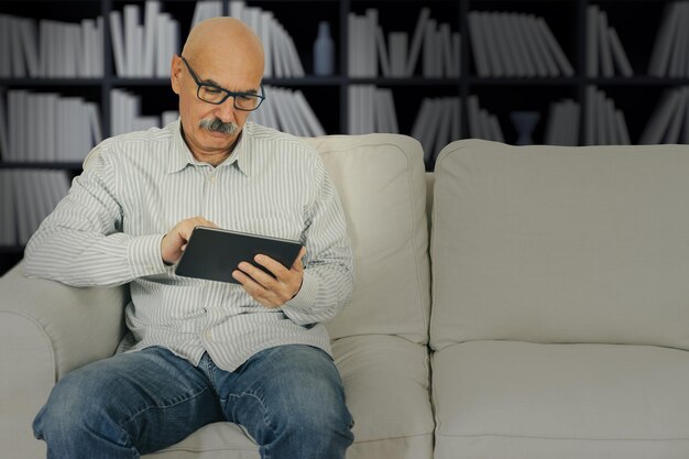 Hombre senior leyendo en una tableta digital sentado en el sofá en la sala de estar de la casa