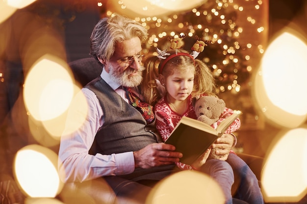 Hombre senior de moda alegre con cabello gris y barba sentado con niña en el libro de lectura de la sala de Navidad decorada.