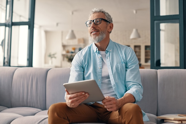Hombre senior pensativo con tableta digital y mirando a otro lado con una sonrisa mientras está sentado en el sofá en casa
