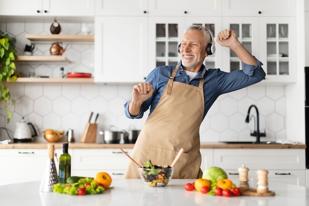 Hombre senior positivo en auriculares escuchando música mientras cocina comida en la cocina