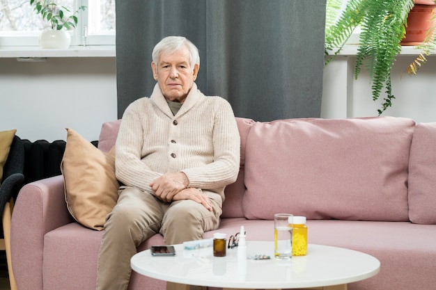 Hombre senior en ropa casual sentado en el sofá junto a una mesa pequeña con pastillas y un vaso de agua y esperando al médico contra las ventanas de la sala de estar
