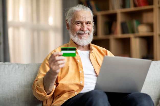 Hombre senior sonriente usando laptop y tarjeta de crédito en casa