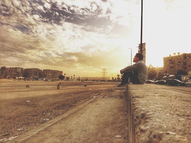 Foto hombre sentado en la acera por la carretera en la ciudad contra el cielo