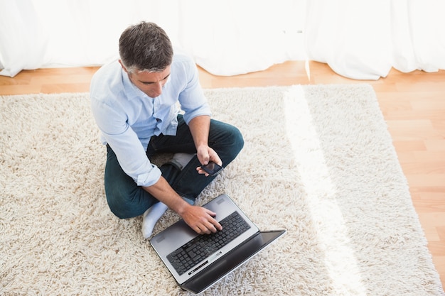 Hombre sentado en la alfombra usando la computadora portátil y el teléfono