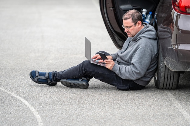 Hombre sentado en el asfalto en automóvil y trabajando en una computadora portátil que trabaja de forma remota viajando