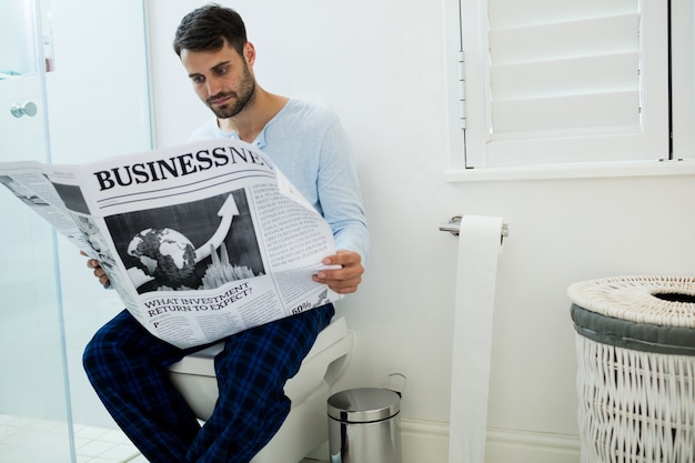 Hombre sentado en el asiento del inodoro leyendo un periódico en casa