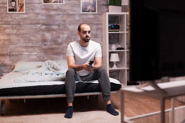 Hombre sentado en el borde de su cama por la noche viendo la televisión con control remoto.