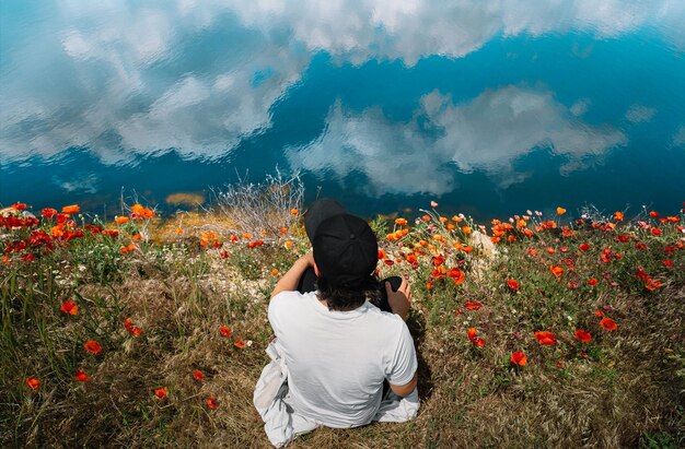 Foto hombre sentado en el campo de amapola junto al lago con el reflejo de las nubes