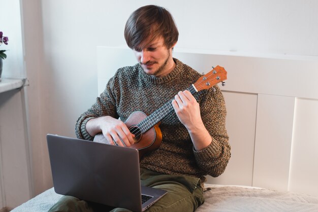 El hombre sentado en casa aprende a tocar el ukelele usando lecciones en línea.